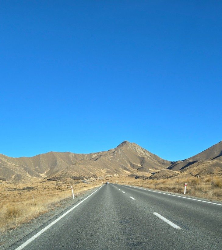 Driving through Lindis Pass, an empty highway running between bare golden tussock hills under a clear blue sky