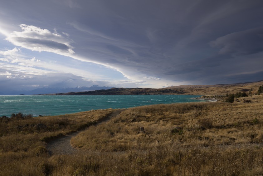The turquoise water of Lake Pukaki under heavy cloud, with a walking track winding away through golden tussock