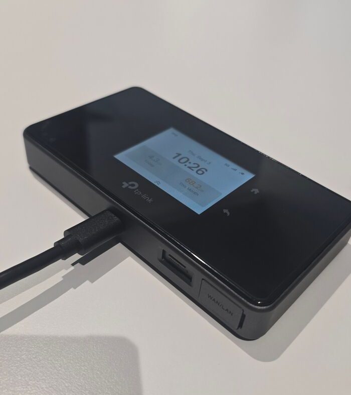 TP-Link M8550 5G mobile router on a white desk, touchscreen showing the clock and data usage for the day and month